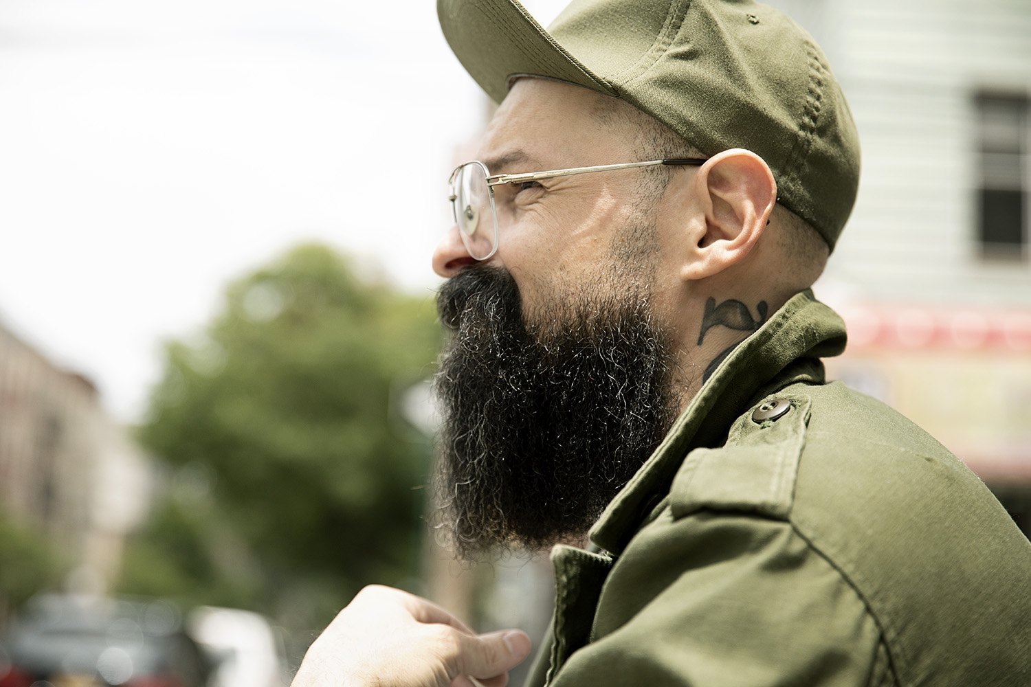 Profile of a bearded man in a green field cap and olive jacket, Brooklyn street soft behind him