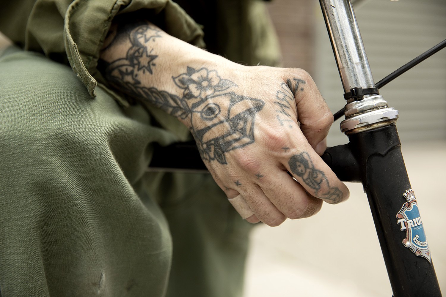 Detail of a heavily tattooed hand gripping the handlebar against the olive sleeve of the jacket