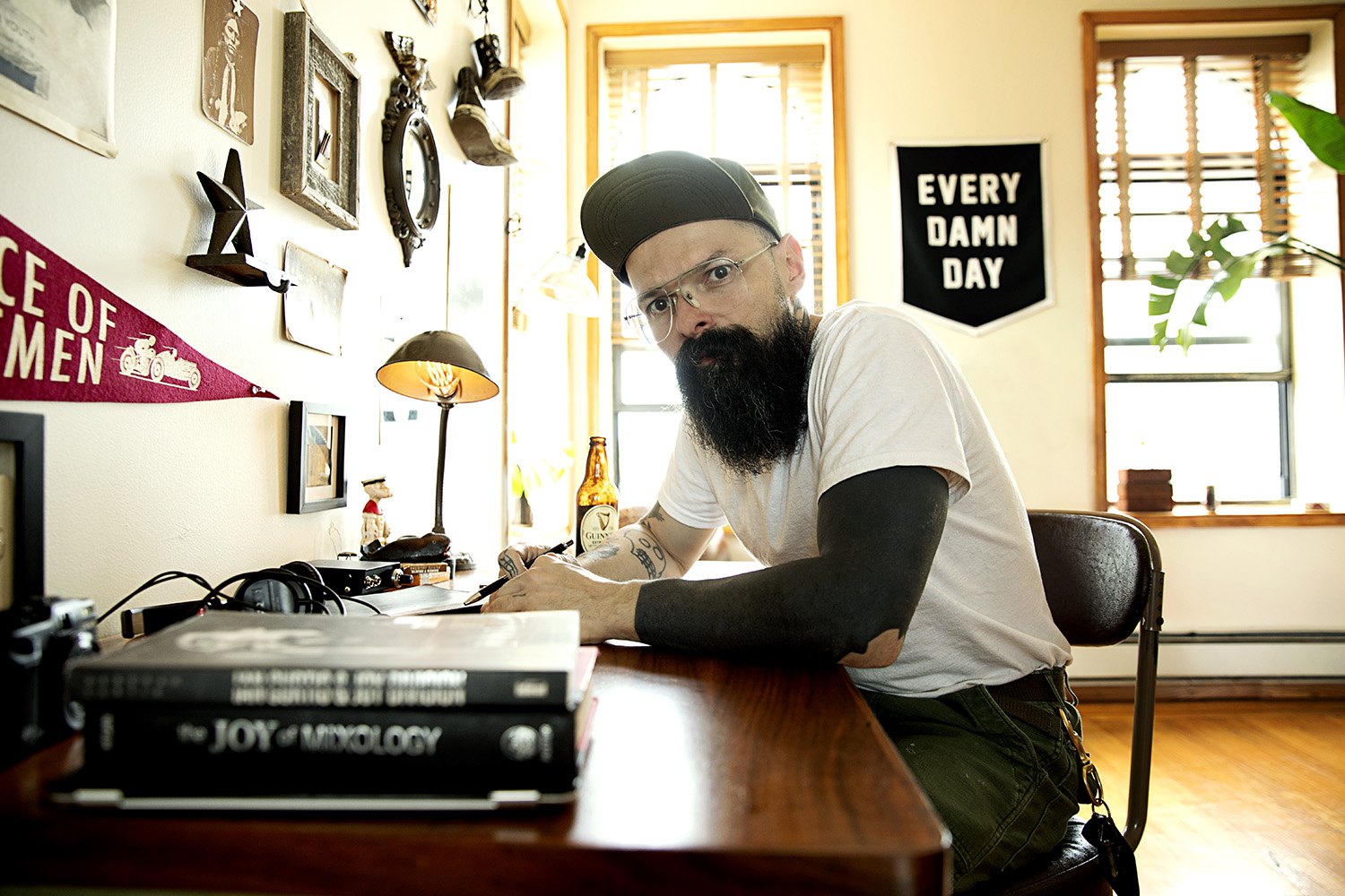 At his desk at home, an EVERY DAMN DAY print on the wall and afternoon light through the window