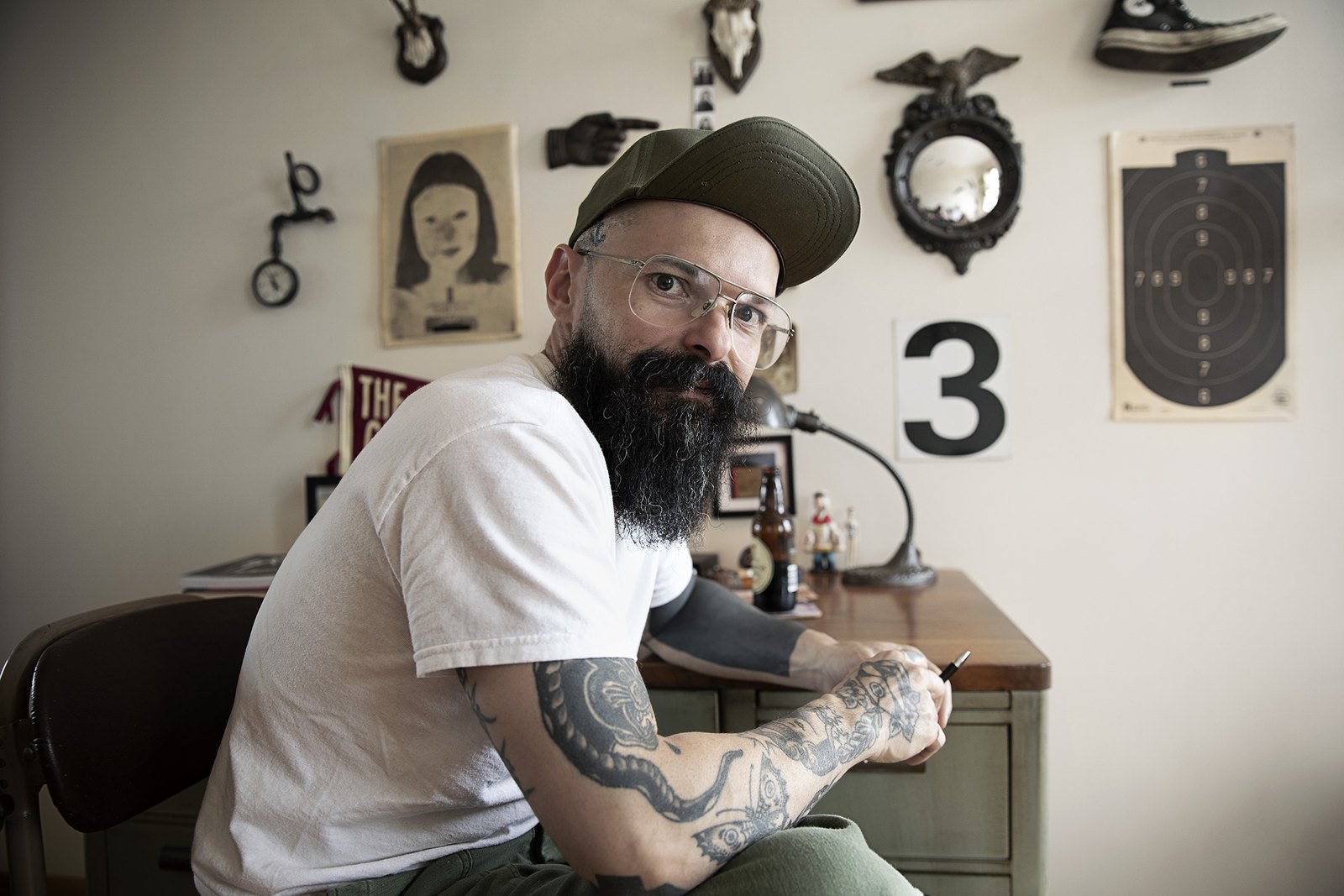 Turning from the desk in a white t-shirt, tattooed forearm resting on the chair back