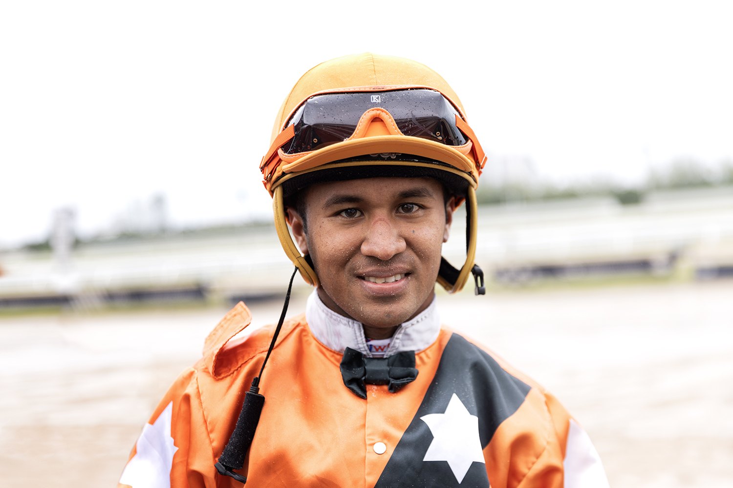 A jockey in orange silks and helmet with a white star on the shoulder, track blurred behind him