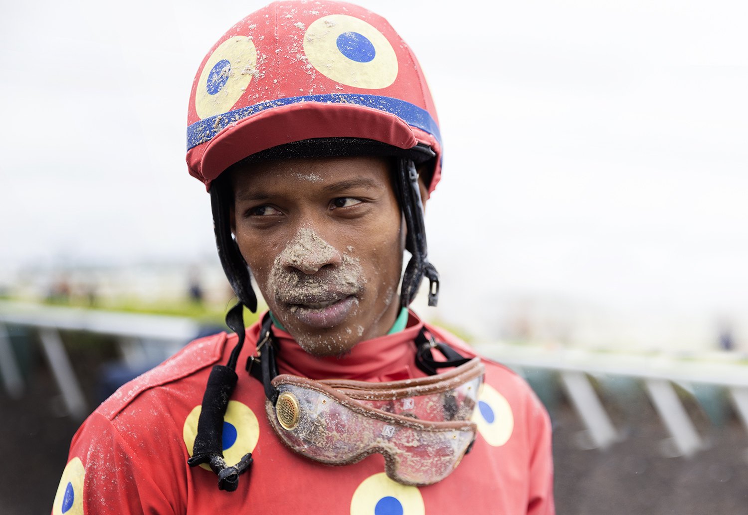 A jockey in red silks with yellow and blue circles, his face still caked in track dirt after the race
