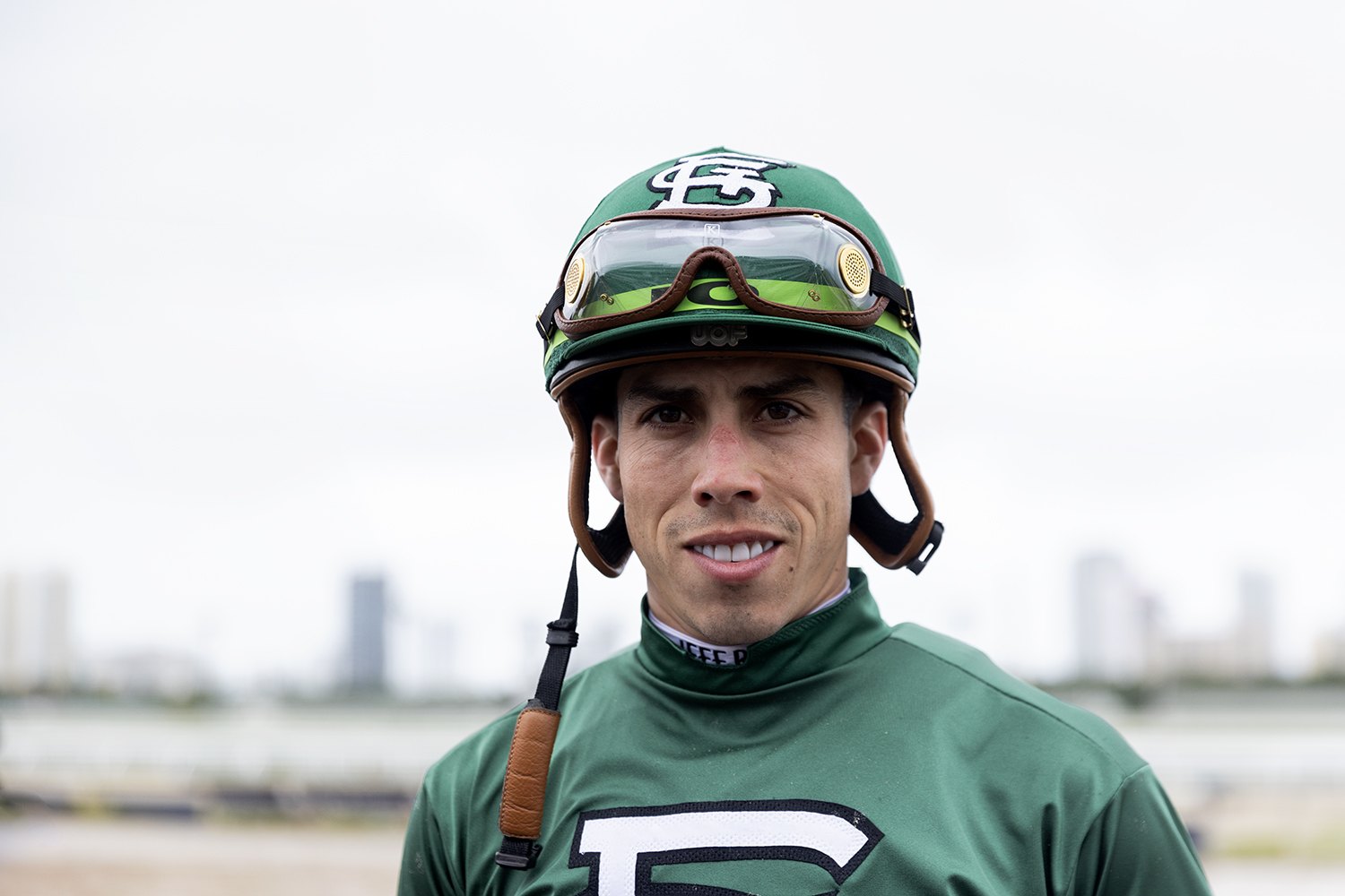 A jockey in green silks and matching helmet looks into the lens, the city skyline behind him