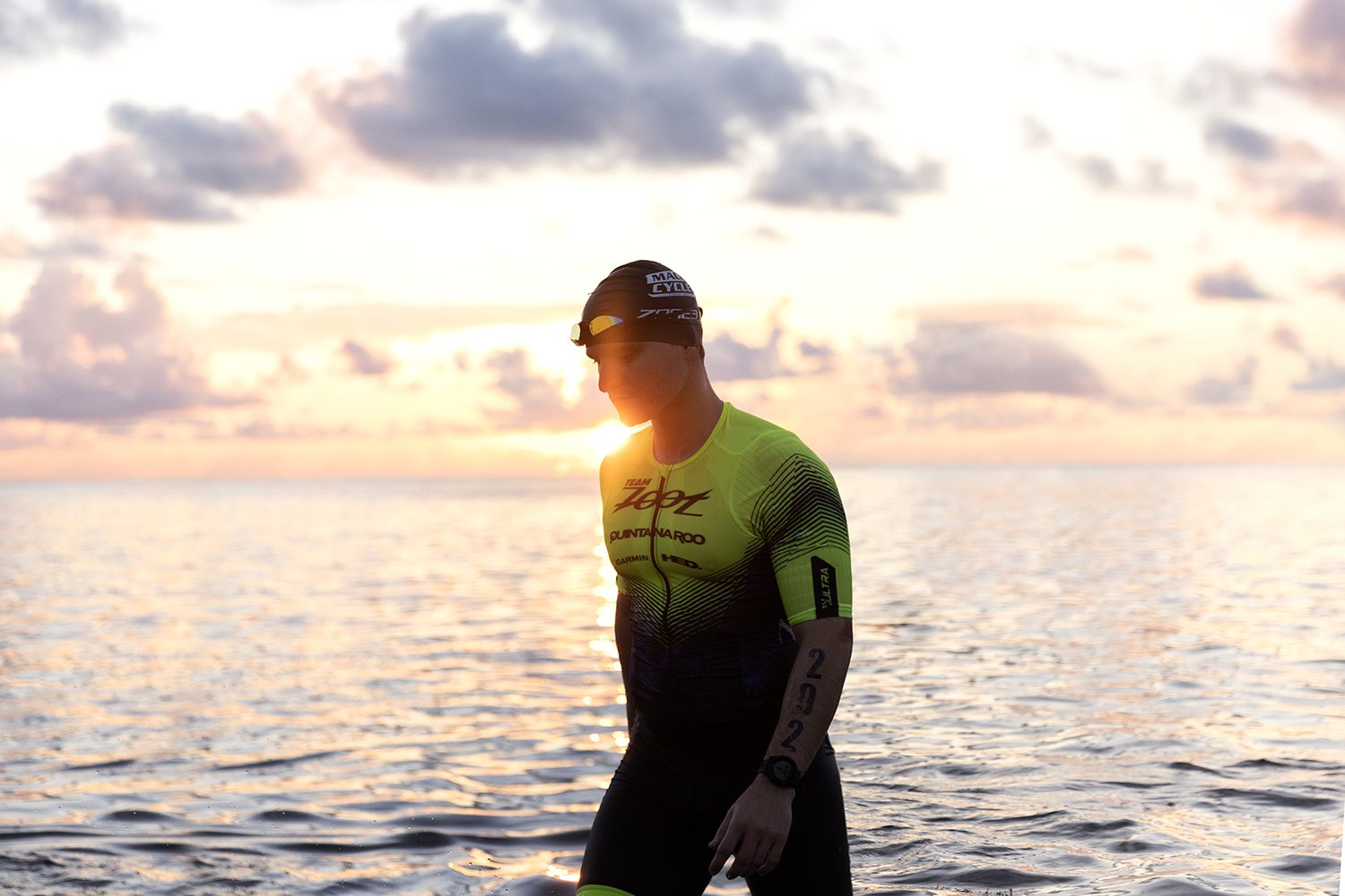 A triathlete in a wetsuit looks out at the sunrise sea