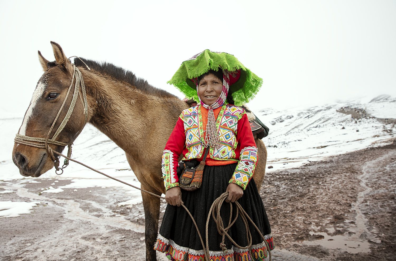 A woman in a green montera stands beside her horse in the snow