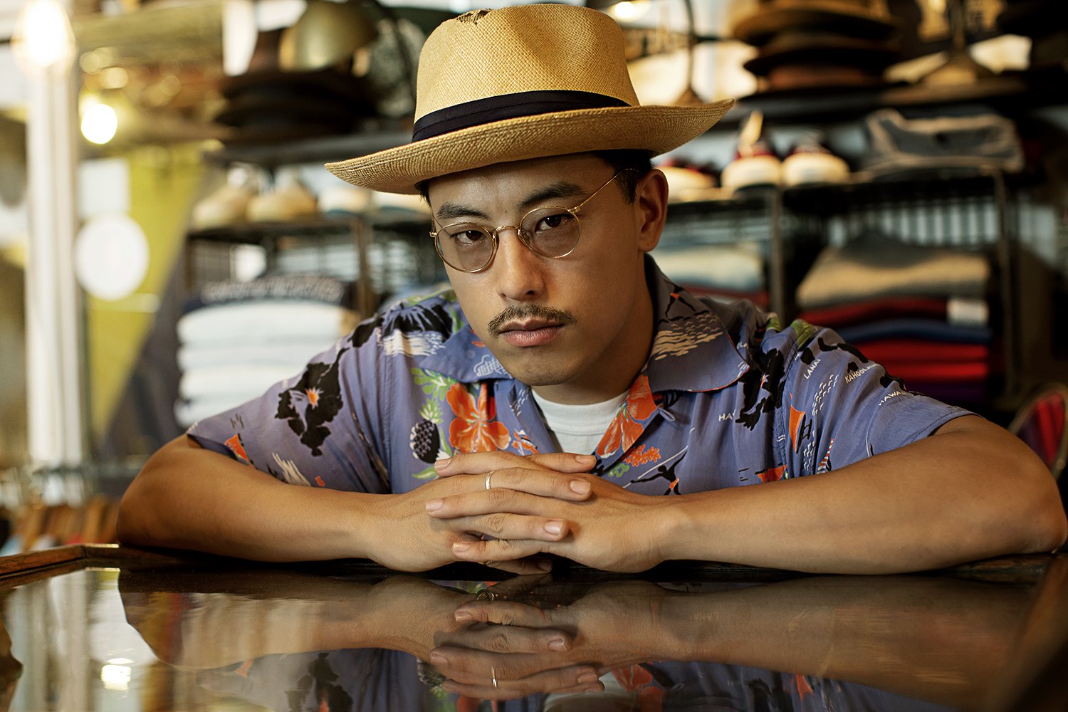 A man in a straw hat and printed shirt leans on a glass counter inside a crowded shop