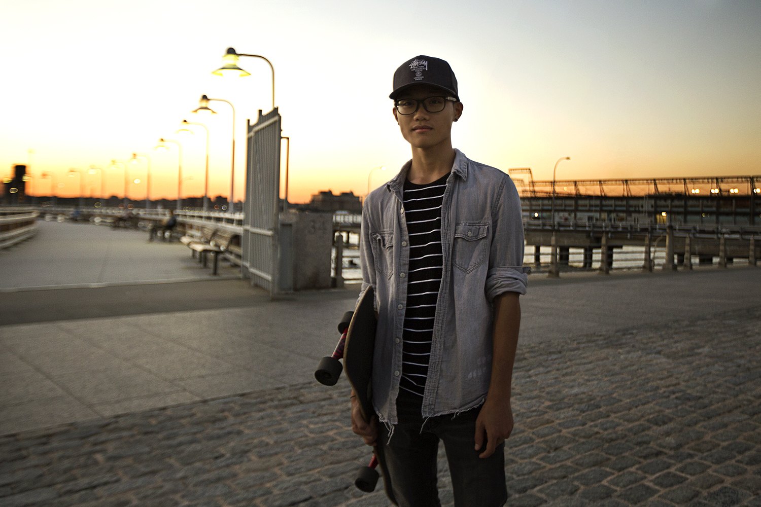 A skater standing with his board at dusk, lamp posts receding down the empty pier