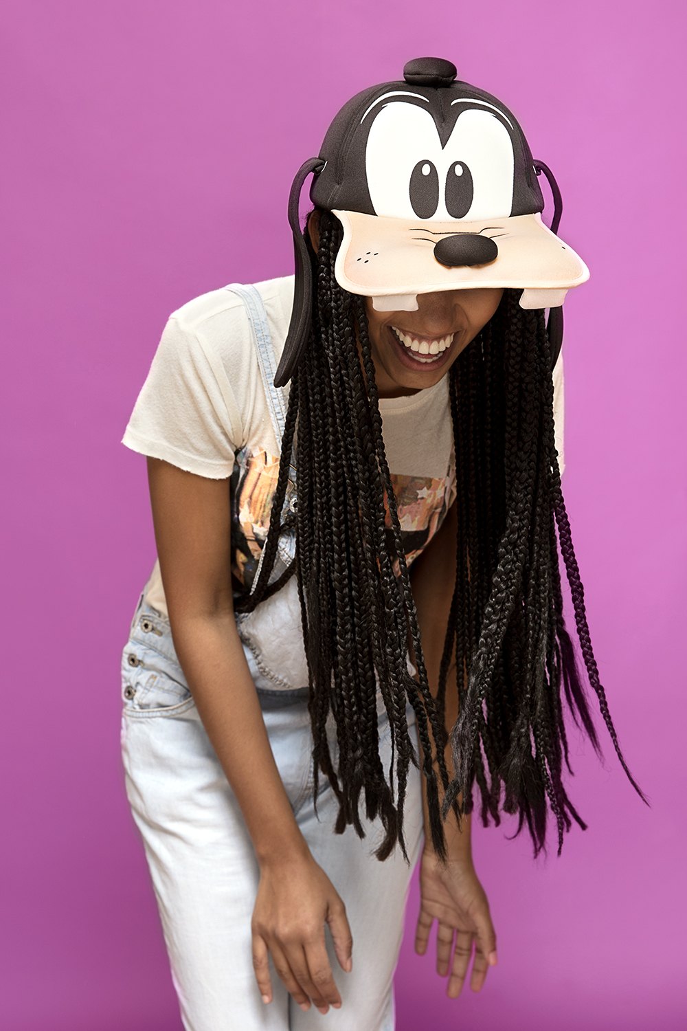 A woman in white dungarees holding a cartoon mask up to her face on magenta