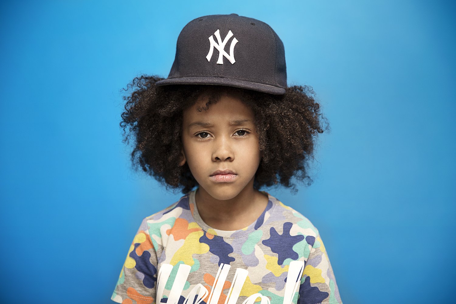 A child in a New York cap and printed shirt against blue