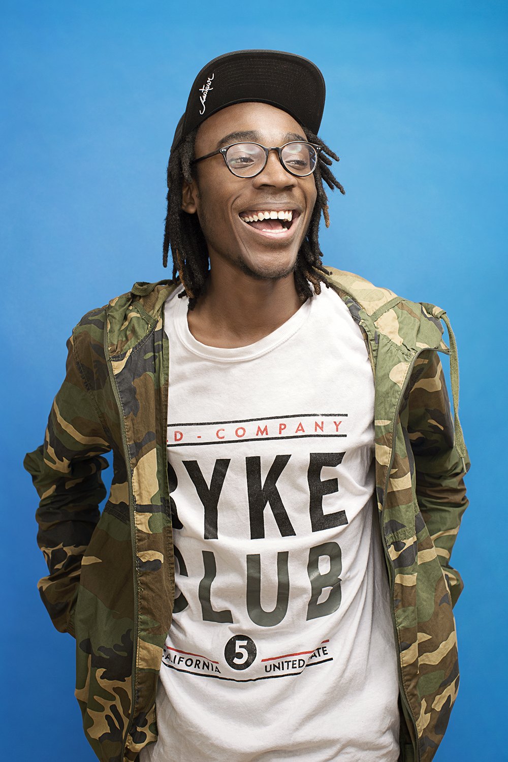 A young man in a camouflage jacket and cap grinning against deep blue