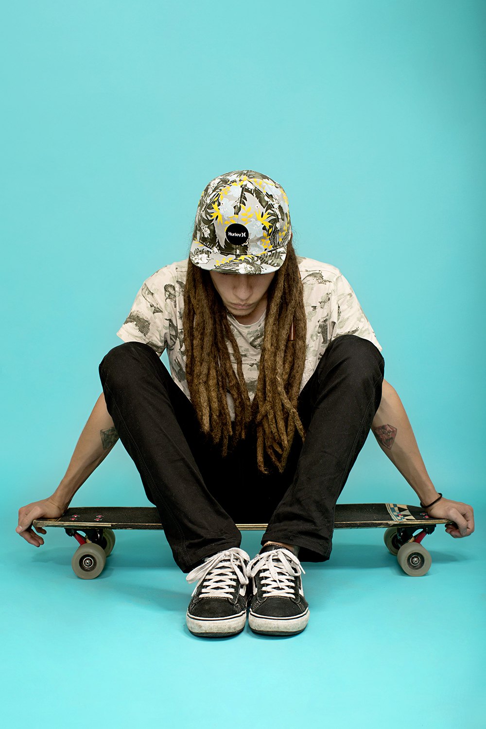 A skater sitting on her board against pale turquoise, hood up