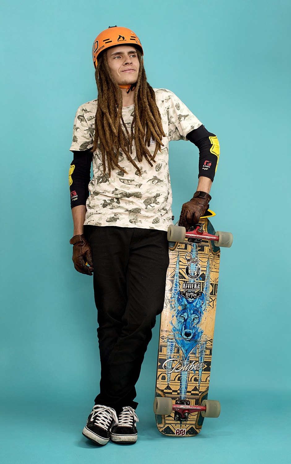 Standing with a patterned skateboard upright at her side, orange cap