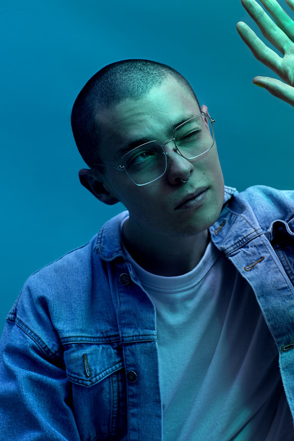 A man in denim under heavy cyan light, hands raised behind him