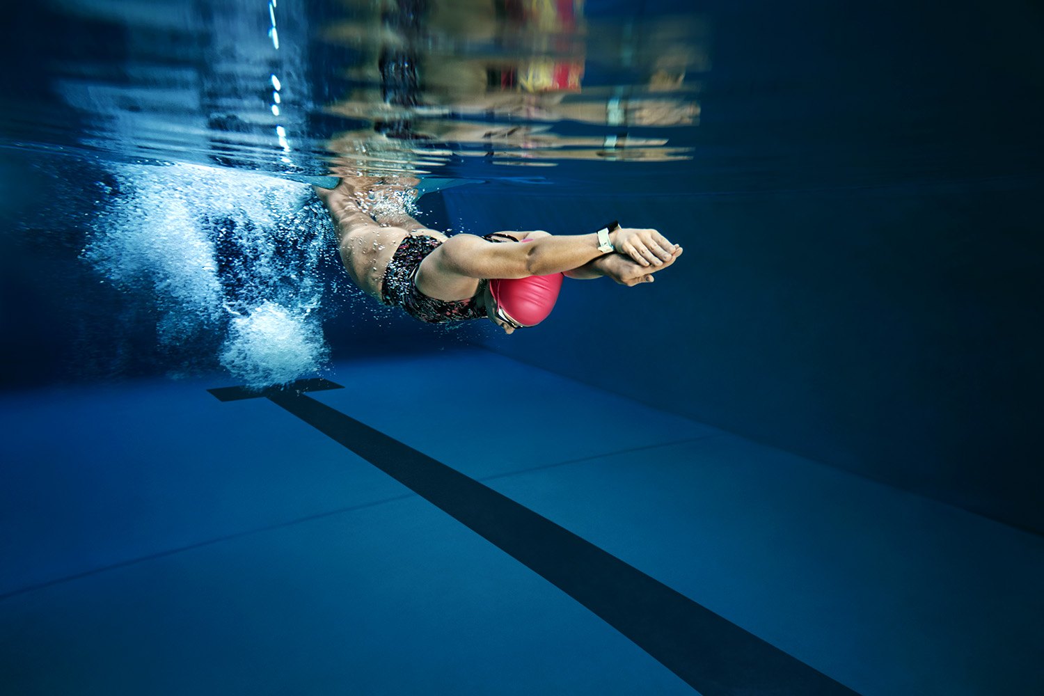 Underwater: a swimmer in a red cap drives forward through a trail of bubbles just below the surface