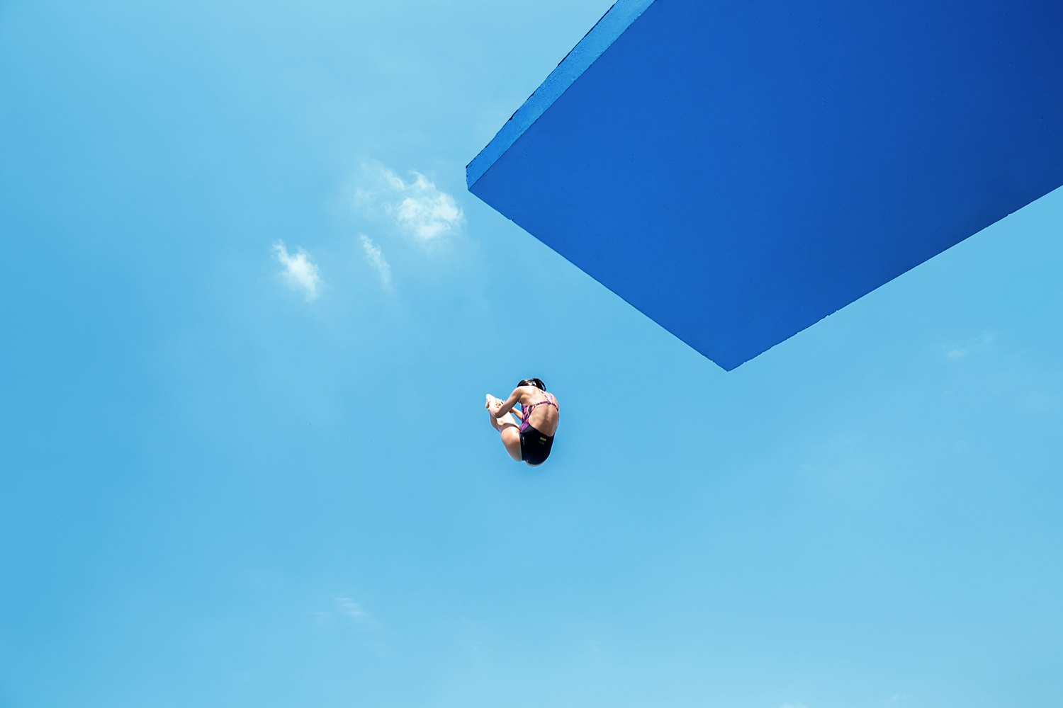 A tucked diver suspended against the sky beneath the platform's blue underside