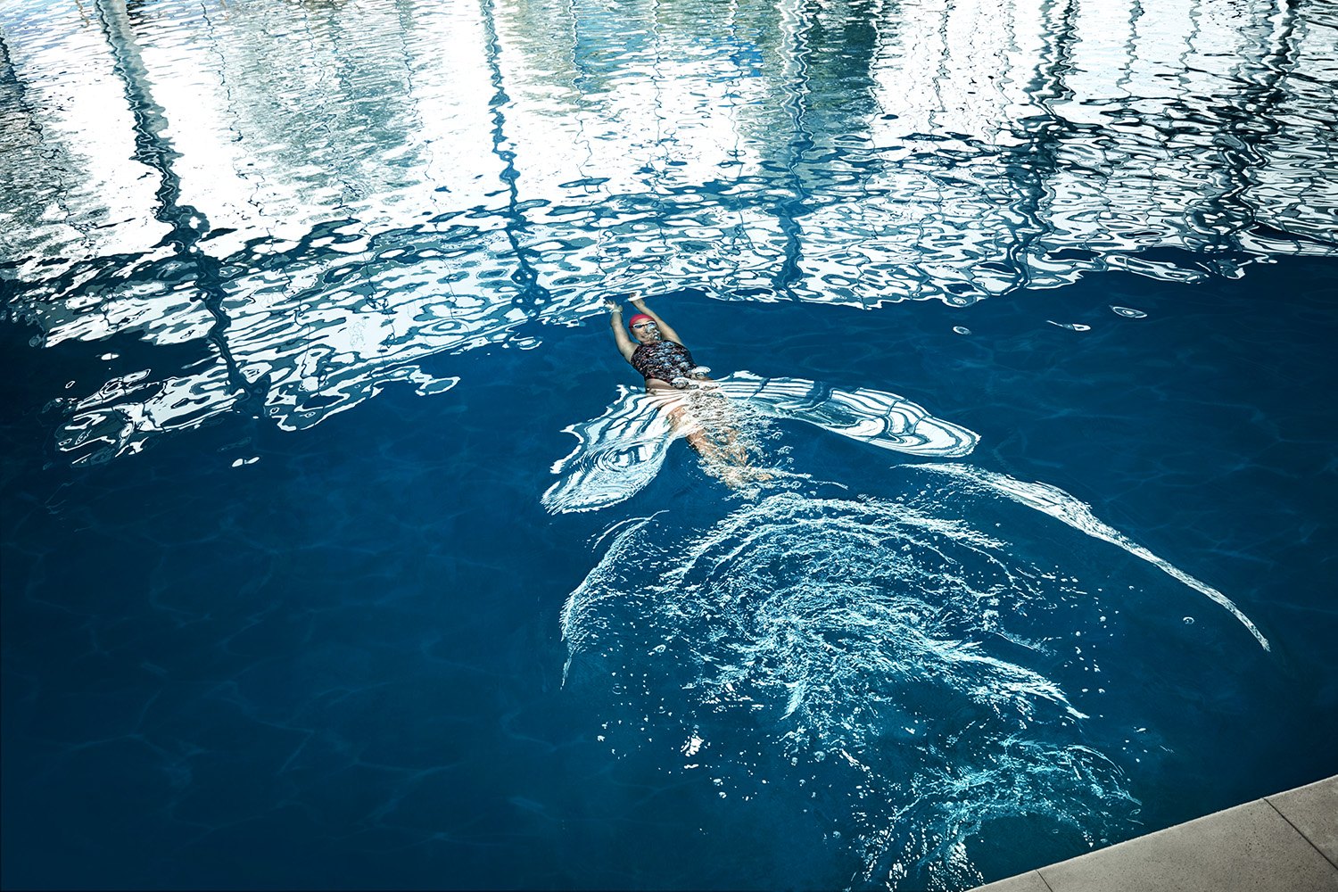 Overhead: a swimmer mid-butterfly in dark water, arms spread, the surface breaking into reflections