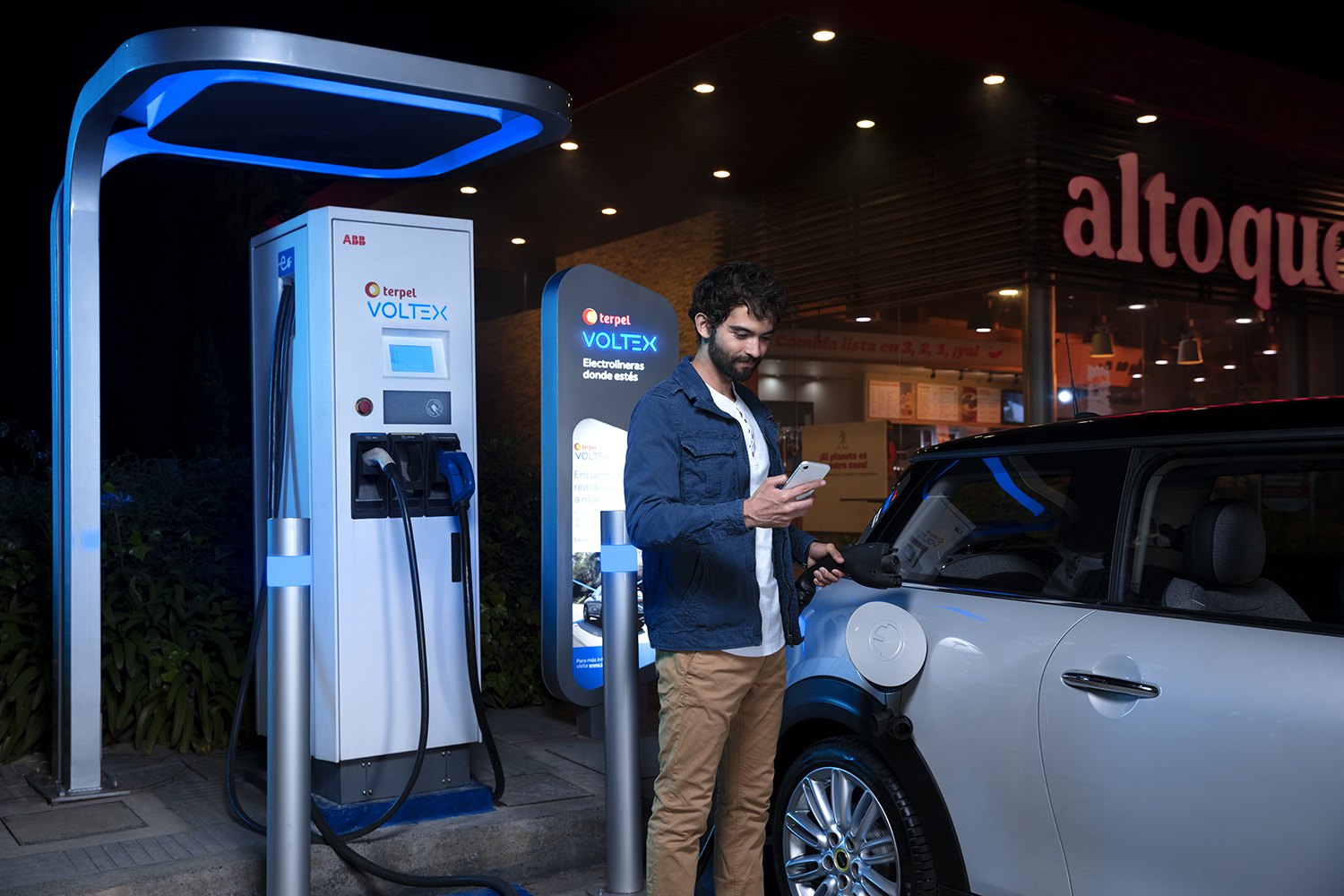 A man checking his phone beside an electric charging bay at a lit service station