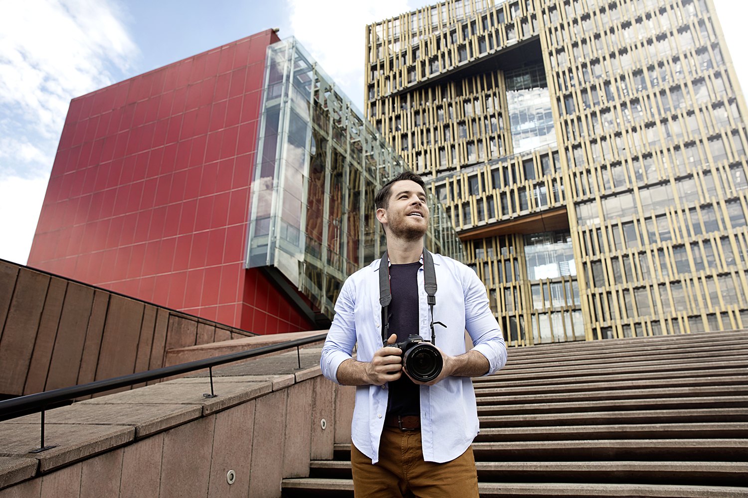 Standing with a camera at the foot of a red and gold contemporary building, broad steps rising behind