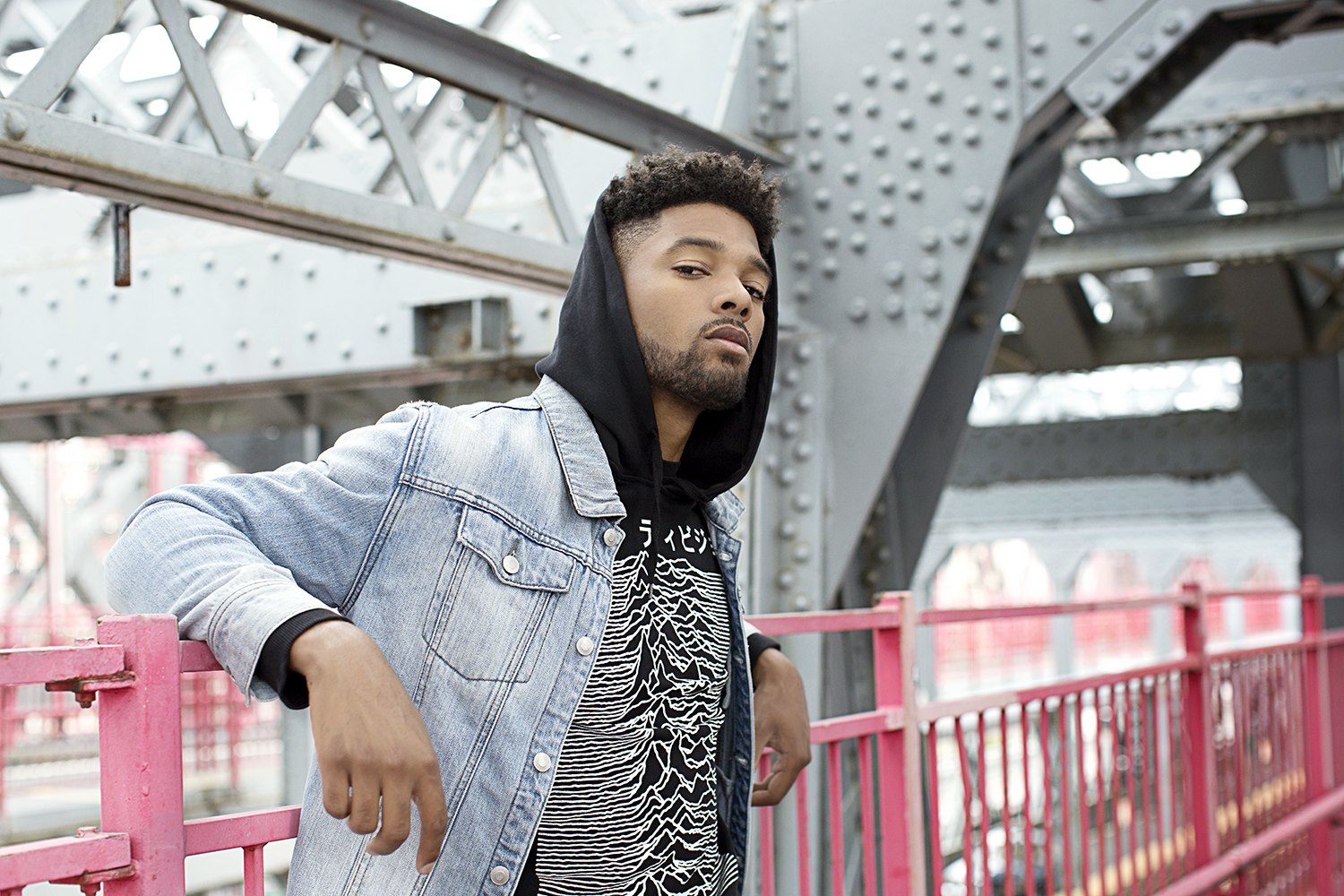 A man in a denim jacket and hood leans on the red railing of the bridge walkway