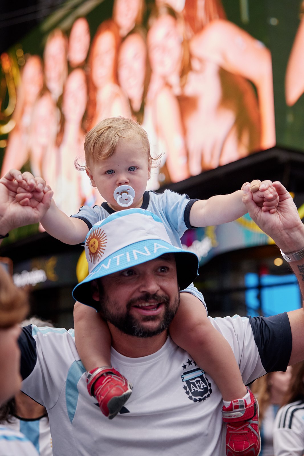 A blond toddler in an Argentina bucket hat sits on his father's shoulders above the march