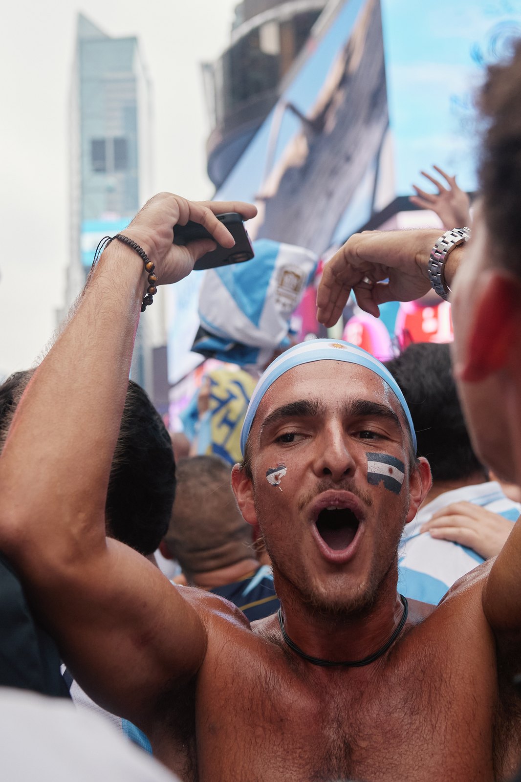 A supporter with an Argentina flag painted on his cheek shouts with his phone raised above the crowd